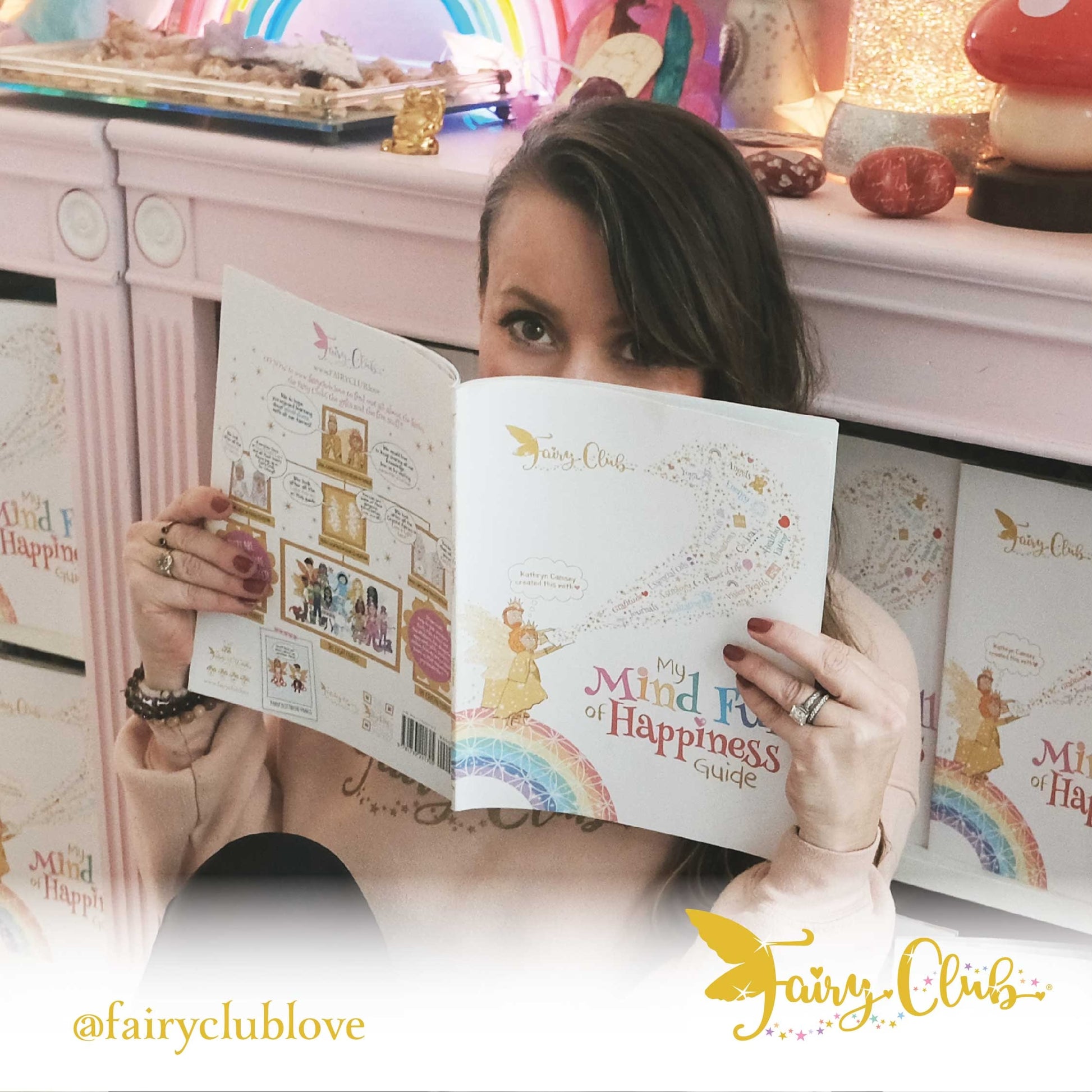 Woman holding a book titled 'My Mind Full of Happiness Guide' with a colorful background featuring the Fairy Club logo.